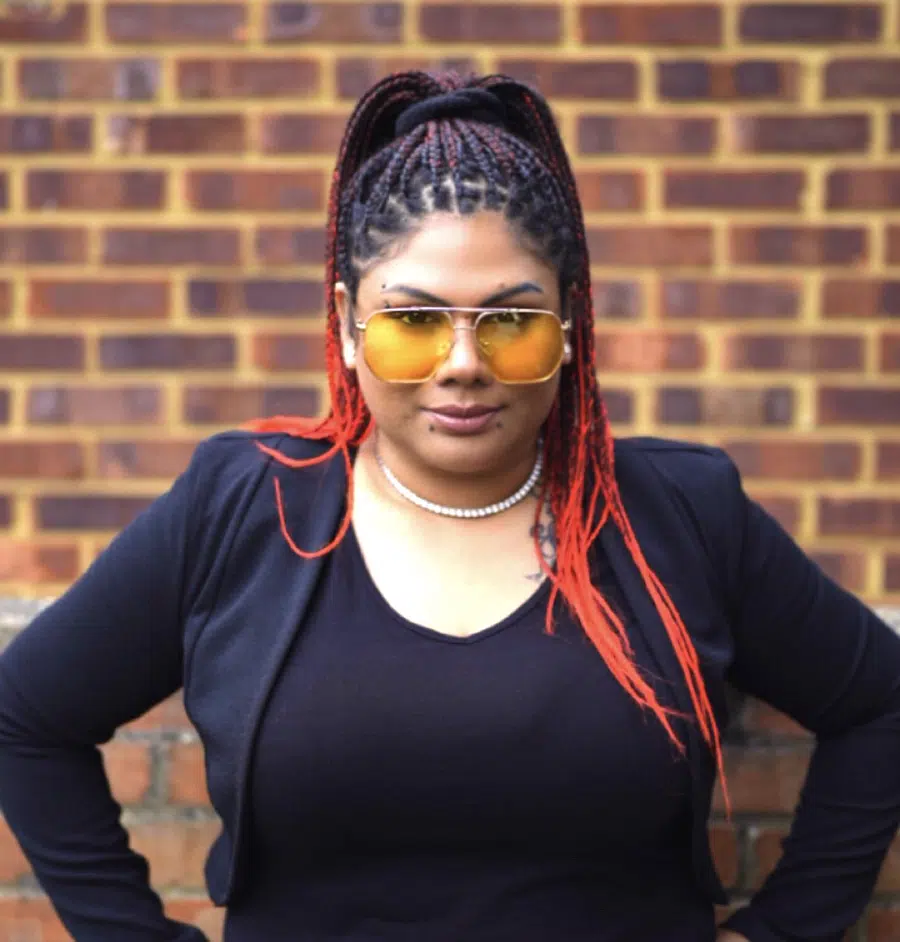 DJ Kim posing against a brick wall wearing yellow sunglasses